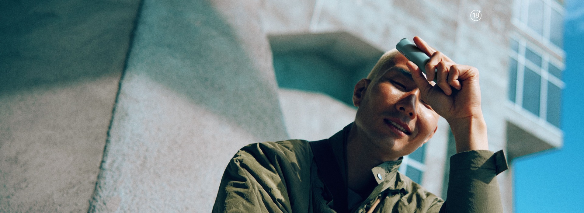 Adult person wearing a green jacket holding an IQOS device outdoors near a modern concrete building under clear blue sky.