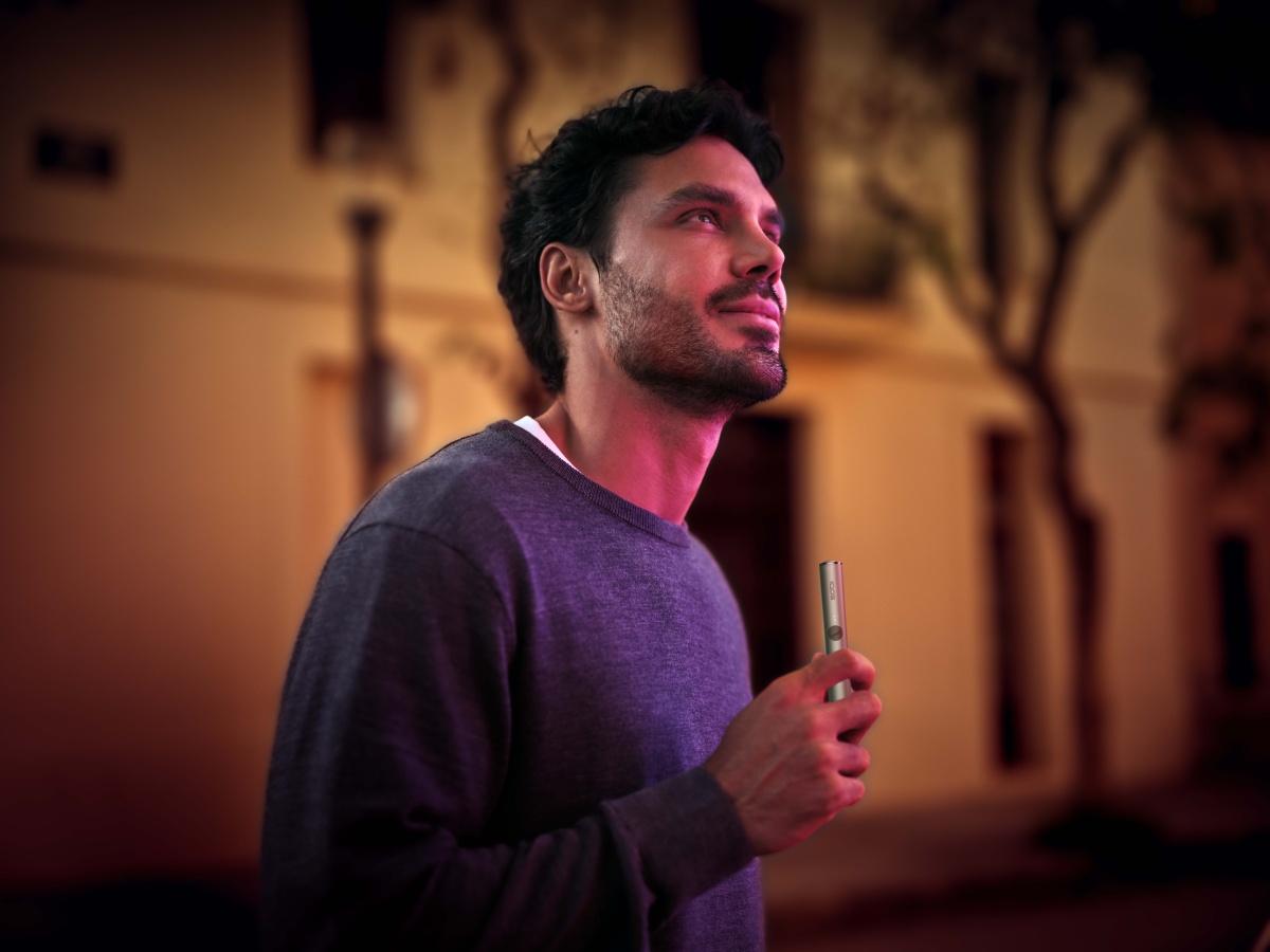 A person standing outside at night, holding an IQOS heated tobacco device. The background shows a blurred building and trees.