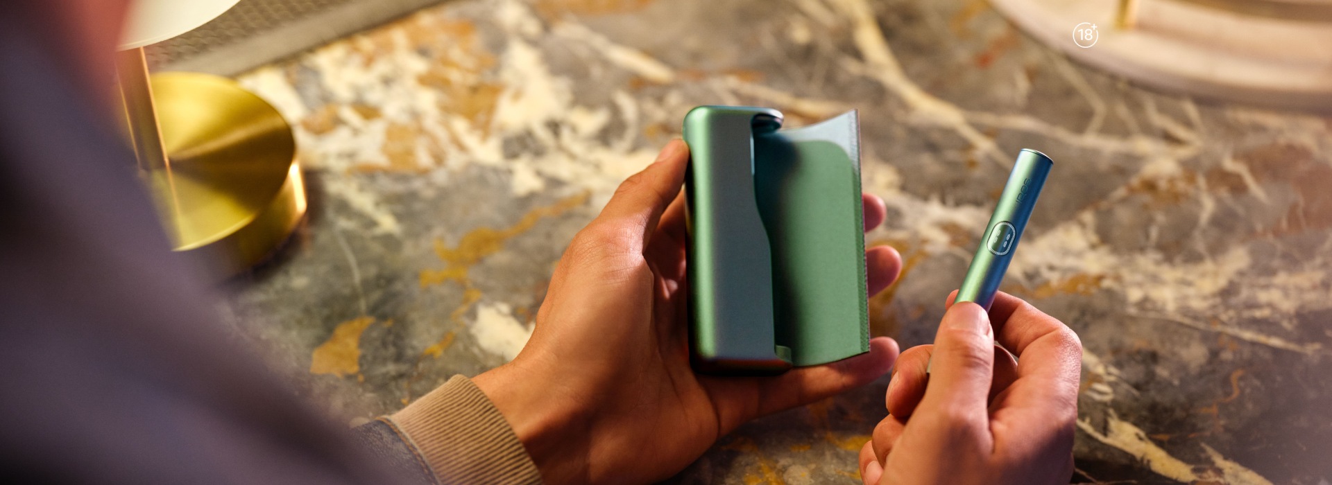 Hands holding IQOS ILUMA i Prime heated tobacco device and charger in Aspen Green on a textured surface.