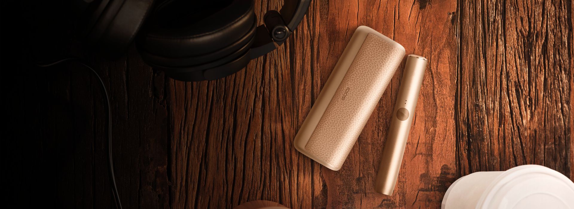 A wooden surface with a pair of black headphones, a beige-colored heated tobacco device, and its case.