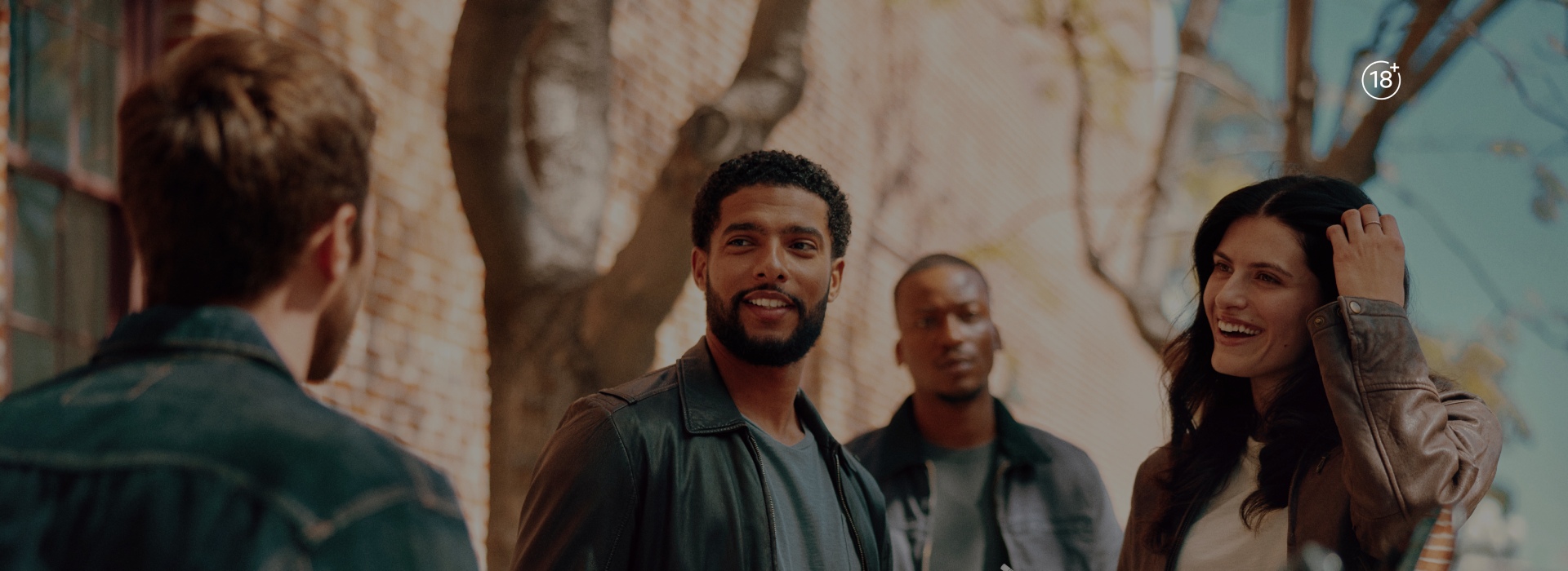 Group of adult people standing outdoors near a brick wall and tree, wearing casual jackets in a sunlit setting.