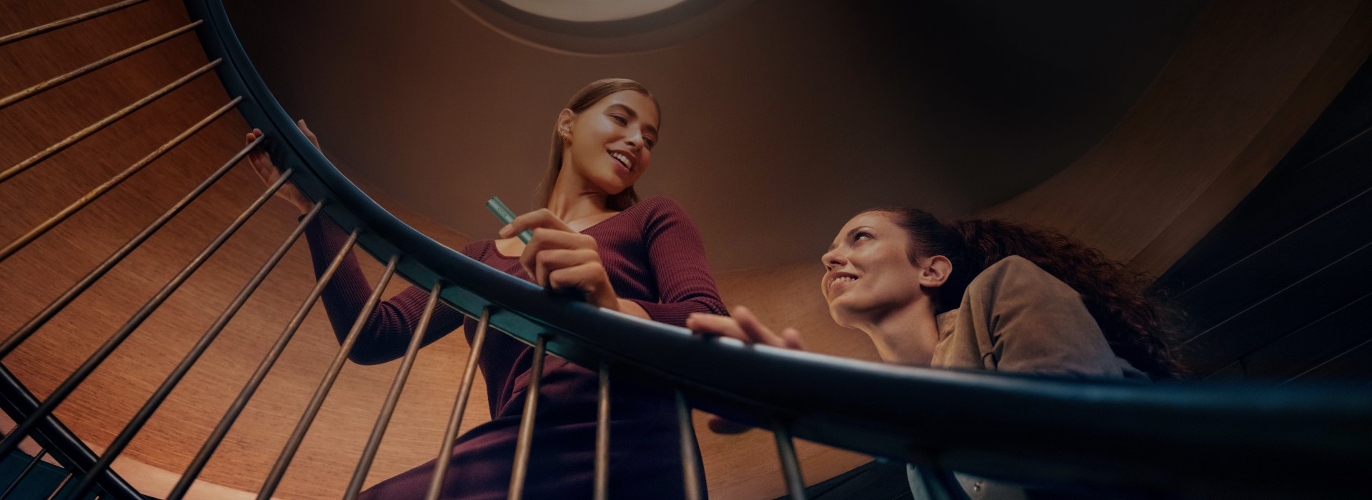 Two people are ascending a spiral staircase. The image is taken from below, looking up towards the ceiling where a circular light is visible. One person is holding onto the railing while the other looks upwards.