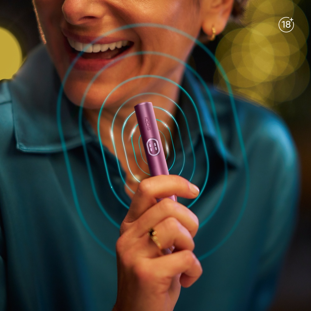 Close-up of a person holding an IQOS ILUMA i PRIME Garnet Red Holder with blue concentric circles indicating vibration, set against a warm, softly blurred background.