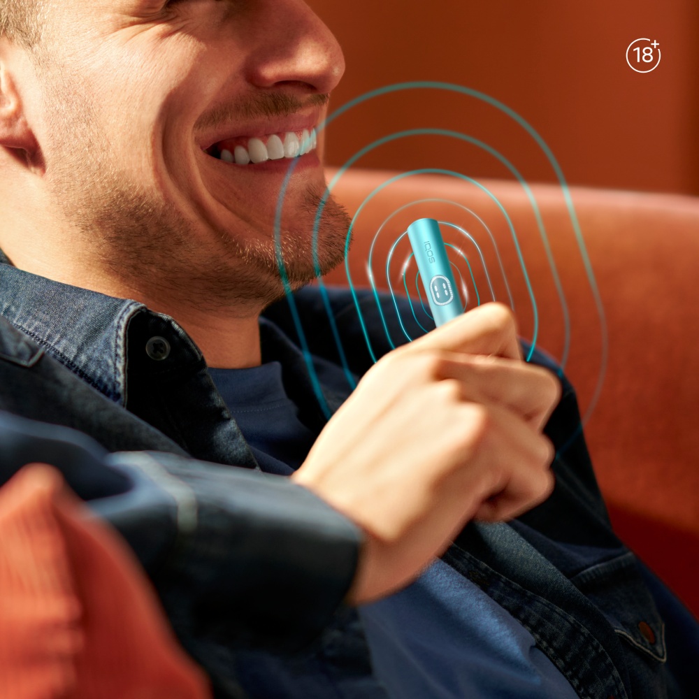 Person holding an IQOS ILUMA i PRIME Breeze Blue Holder emitting blue concentric circles near their mouth, with a blurred background.