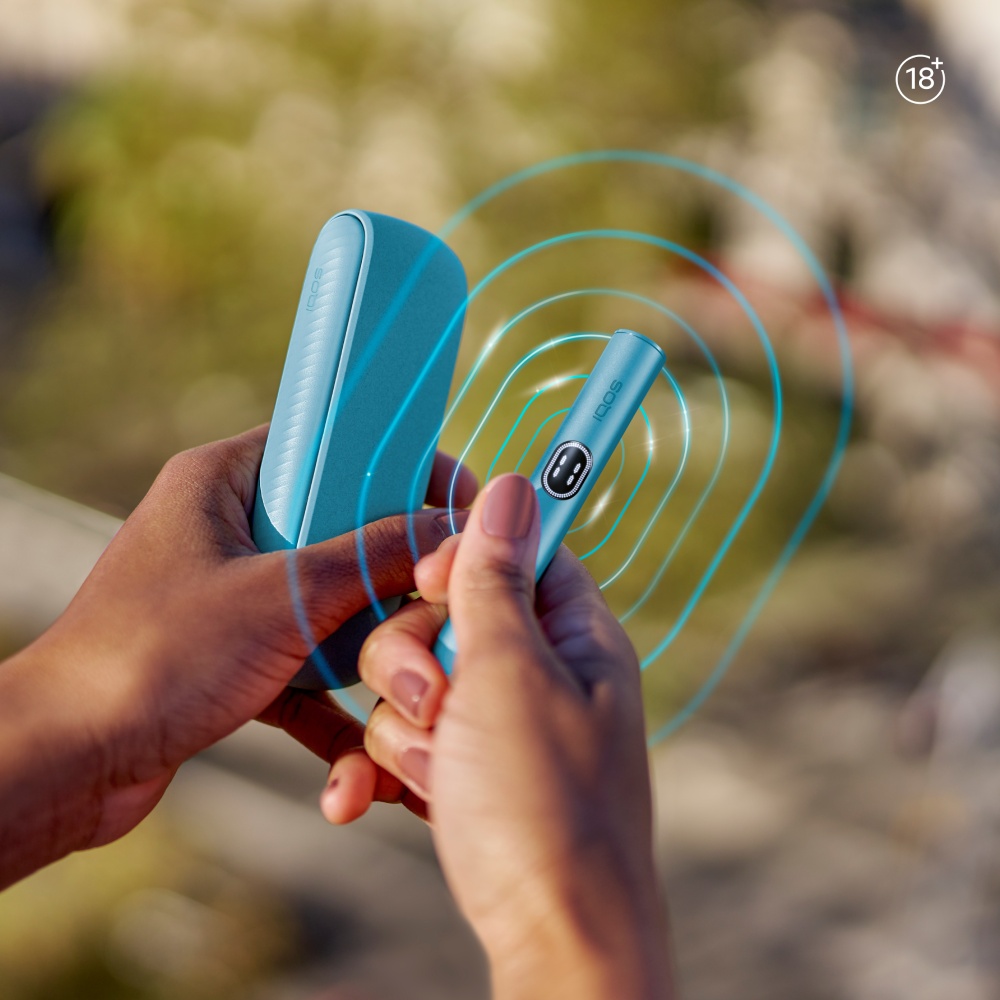 An IQOS ILUMA i PRIME Breeze Blue emitting blue concentric circles , resting on a table with a cup and notepad in the background.