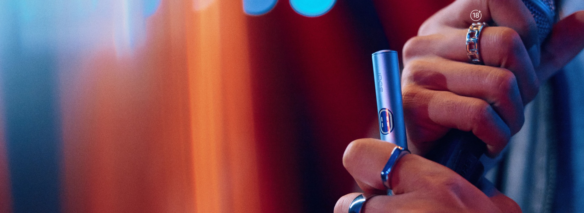 Close-up of hands holding an IQOS ILUMA i heated tobacco device with rings and blurred lights in the background.