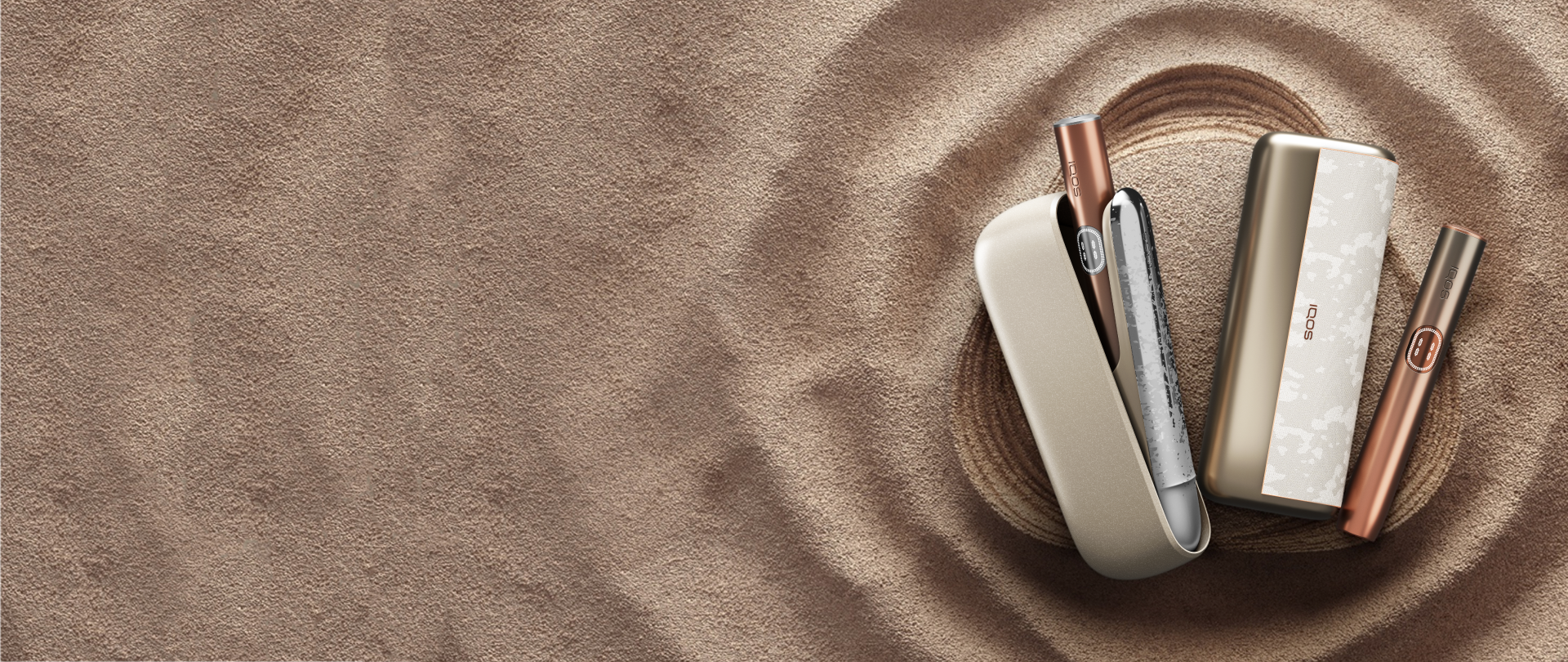 Dve napravi iz serije IQOS ILUMA i MINERA – ILUMA i ter ILUMA PRIME i – ter držala in žepna polnilnika na peščeni podlagi.