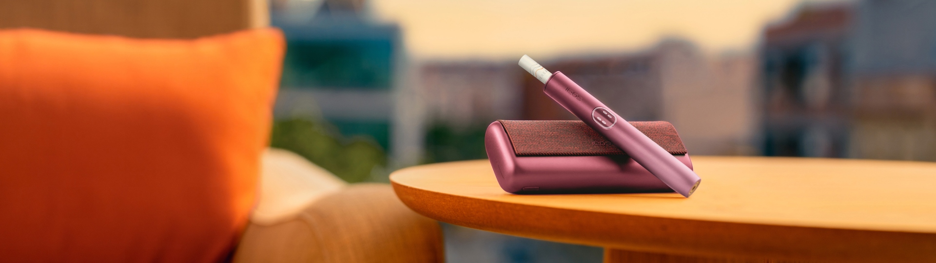 Zařízení IQOS ILUMA i PRIME v barvě Garnet Red nad svou nabíječkou, na stole poblíž gauče