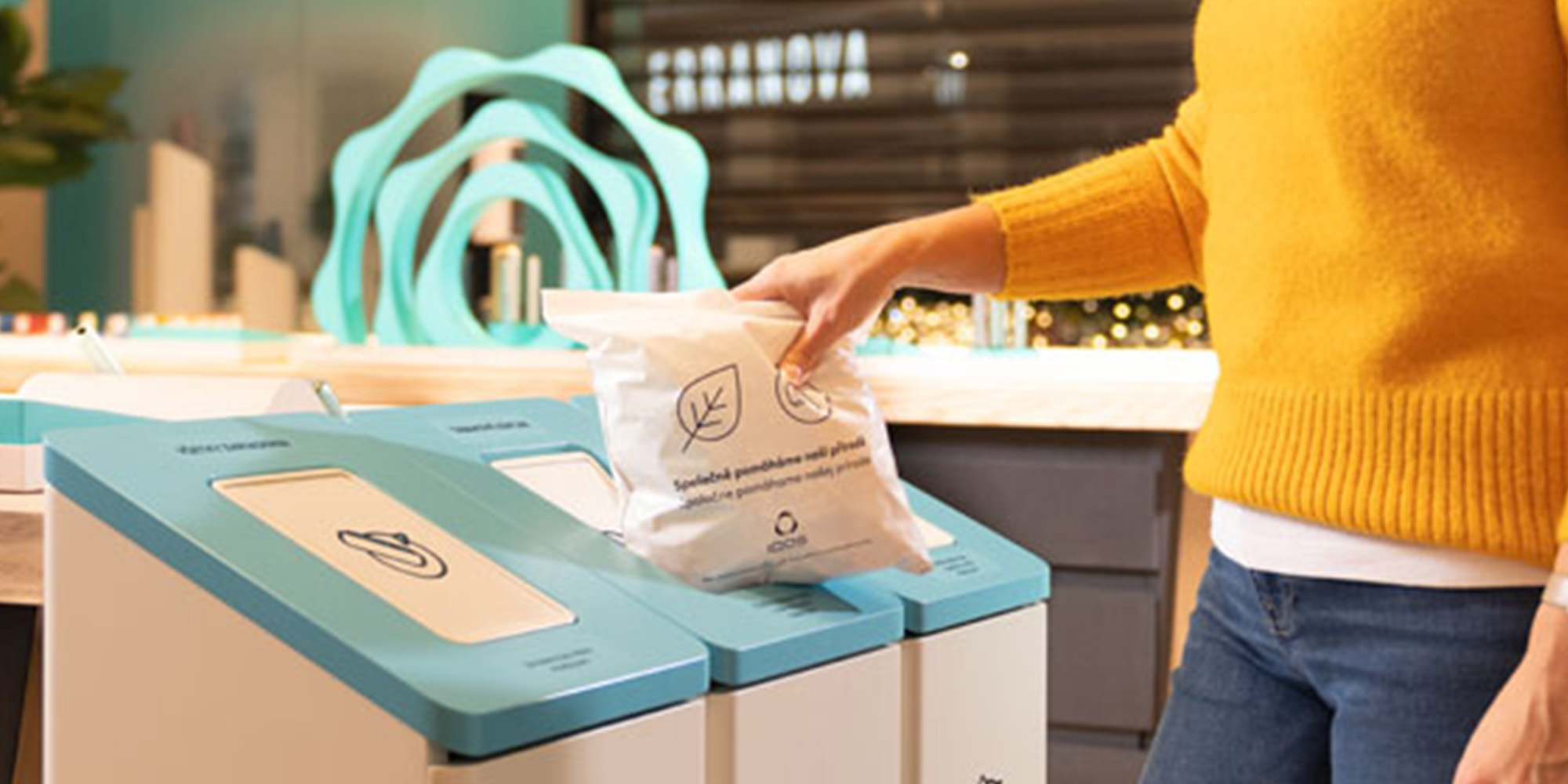 Žena v oranžovom svetri držiaca plný recyklačný sáčok IQOS pri recyklačnom boxe.