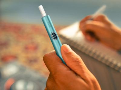 Person holding a Breeze Blue IQOS ILUMA i Prime Holder with backgound of a work environment