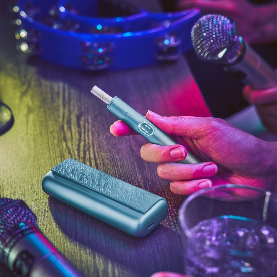 Hand holding a blue IQOS ILUMA i PRIME device; its charger and microphones on a table