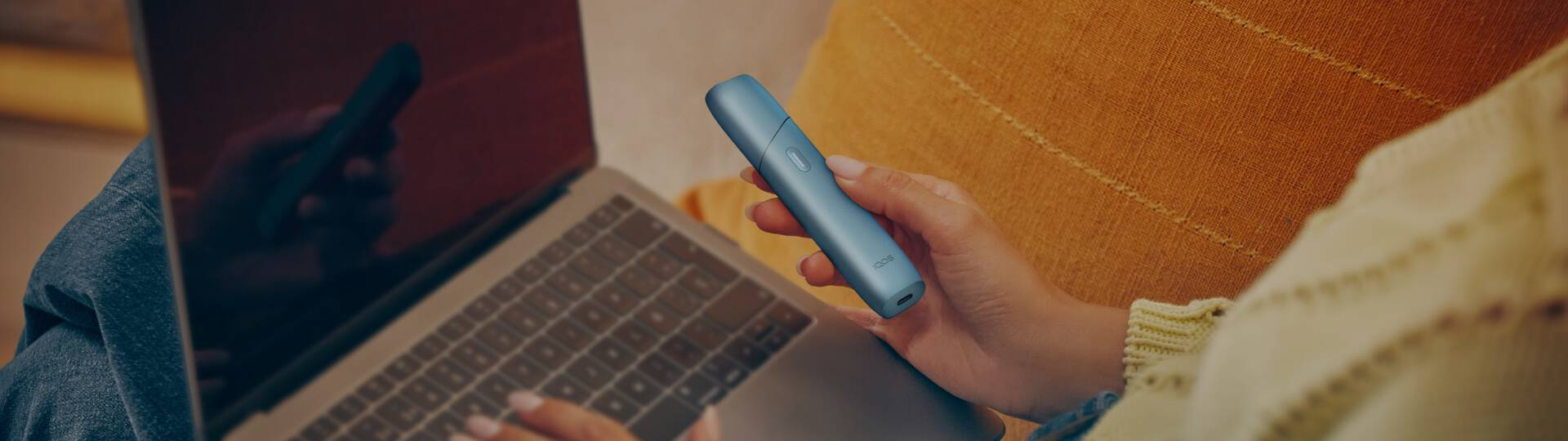 Woman holding an IQOS Originals ONE heated tobacco device and typing on laptop