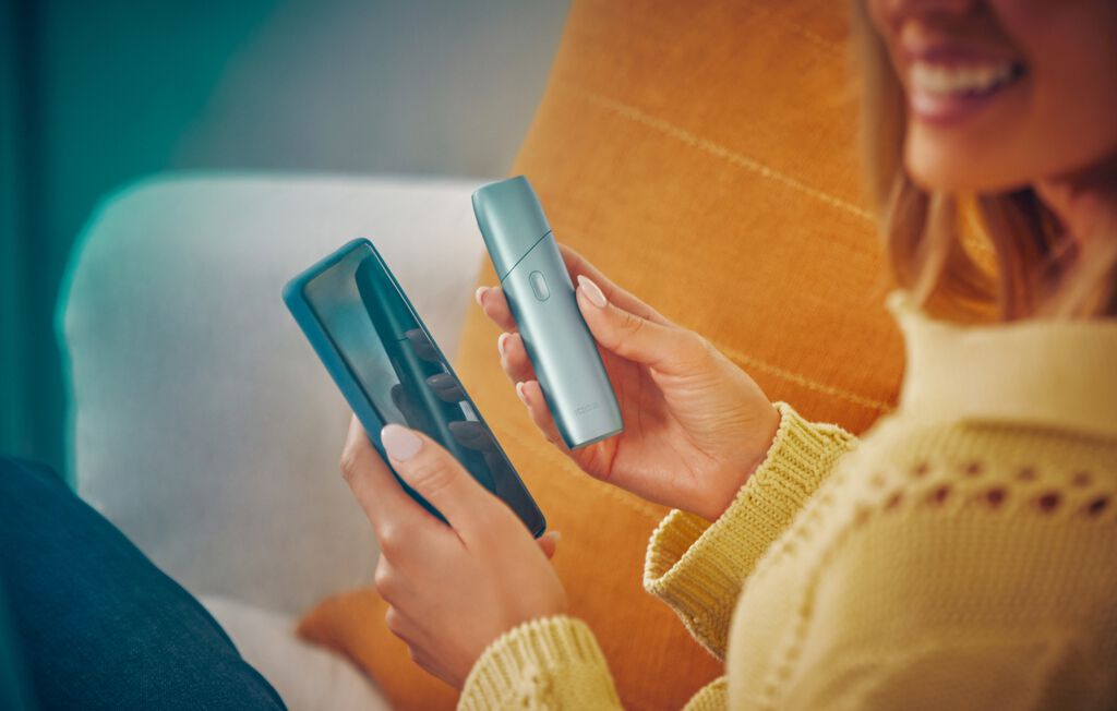 Smiling woman holding a mobile phone and IQOS Originals One device.
