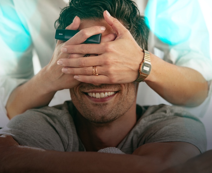 Smiling man with woman's hands hiding his eyes, she's holding a velvet gray IQOS VEEV device