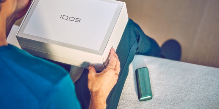 A person holding an unopened IQOS device box. A person holding an unopened IQOS device box.