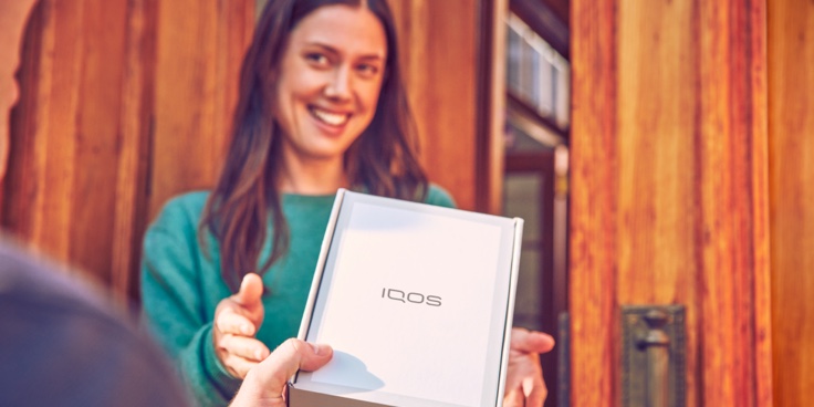 One person handing an IQOS device box to a smiling woman. One person handing an IQOS device box to a smiling woman.