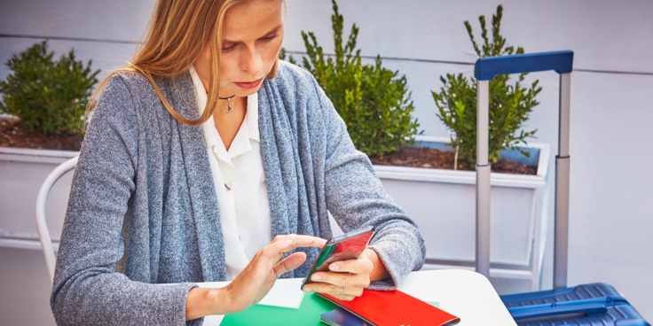 A woman sitting at a table next to her suitcase with a smartphone, passport and plane ticket.