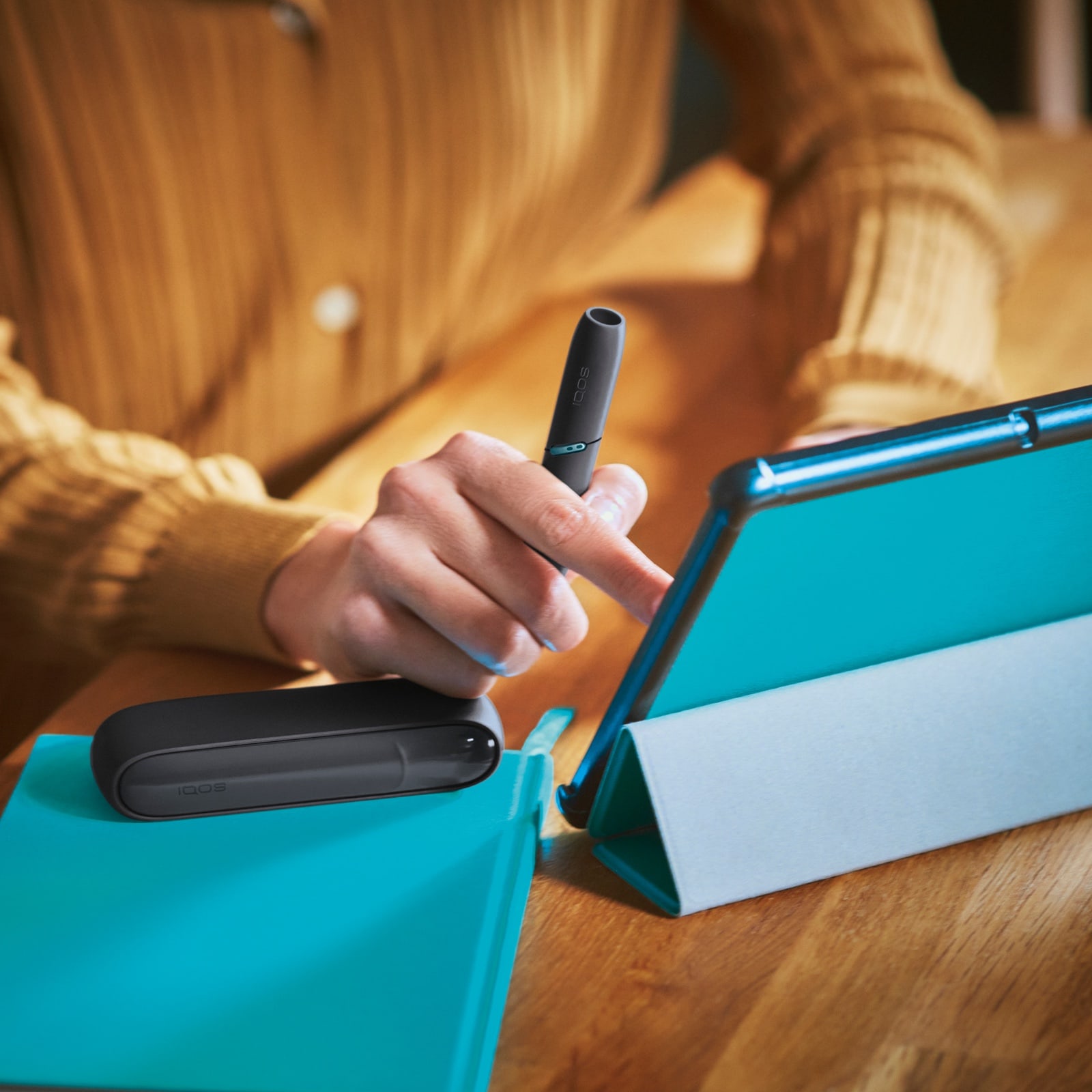 Eine Frau mit orangfarbenem Hemd bedient ein Tablet und hält dabei ein IQOS-Gerät
