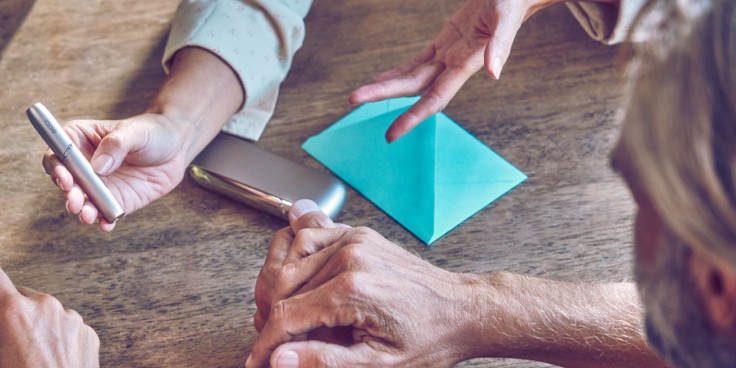 A hand showing an IQOS device to another person.